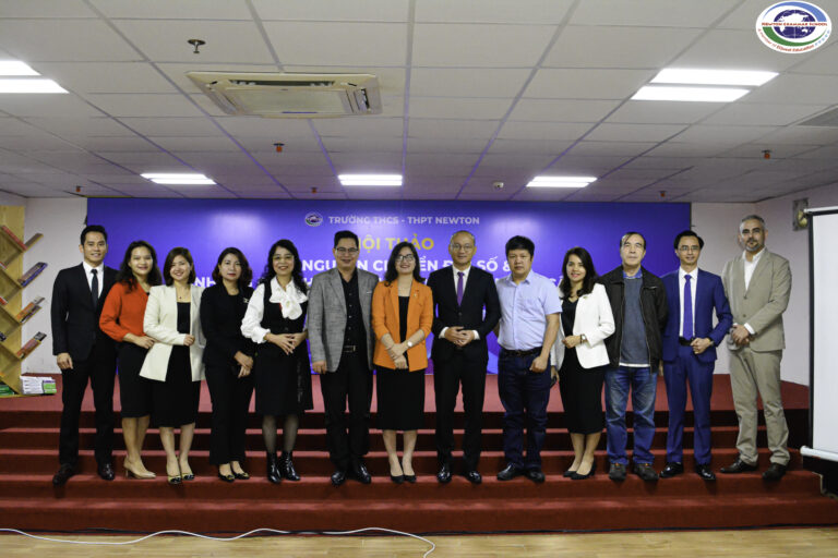 Một nhóm gồm mười ba người, ăn mặc chỉnh tề, đứng sát nhau trên sân khấu có phông nền màu xanh, tạo dáng chụp ảnh tập thể tại một sự kiện hoặc hội nghị.