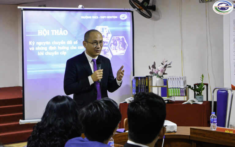 Một người đàn ông mặc vest đang nói vào micro trong một buổi hội thảo. Phía sau anh ta là một màn hình trình chiếu có nội dung tiếng Việt và một bàn có sách và hoa. Một số người đang ngồi và lắng nghe một cách chăm chú.