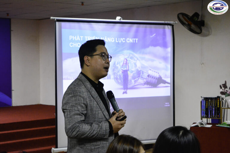Một người đàn ông mặc áo blazer màu xám cầm micro và nói chuyện với khán giả trong một căn phòng. Phía sau anh ta là một slide trình chiếu bằng tiếng Việt về phát triển năng lực công nghệ thông tin, với hình ảnh một phụ nữ kèm theo các biểu tượng công nghệ.