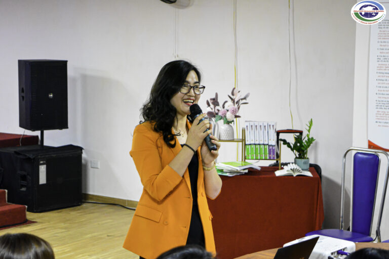 Một phụ nữ mặc áo blazer màu cam mỉm cười và nói vào micro trong một buổi trình bày tại một phòng có bàn màu đỏ, sách và cây cảnh ở phía sau.