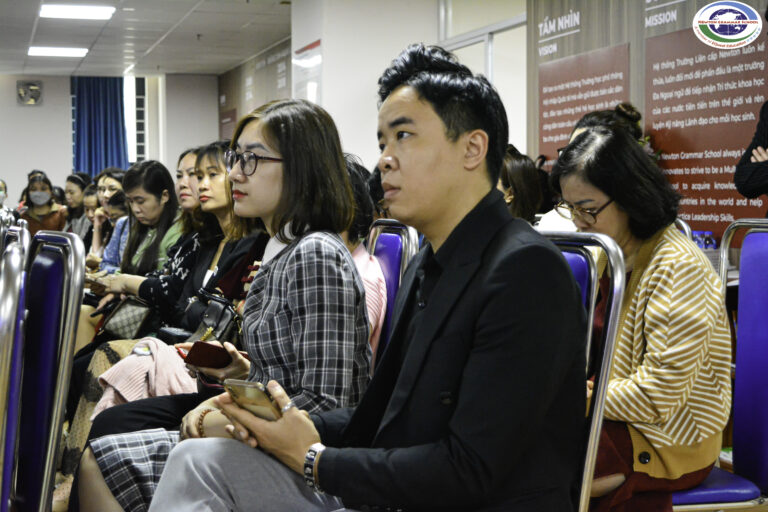 Một nhóm người, chủ yếu là phụ nữ, ngồi thành hàng trên những chiếc ghế màu xanh tại một sự kiện trong nhà. Một số người đang lắng nghe chăm chú, trong khi những người khác nhìn xuống thiết bị của mình. Văn bản tiếng Việt có thể nhìn thấy trên tường ở phía sau.