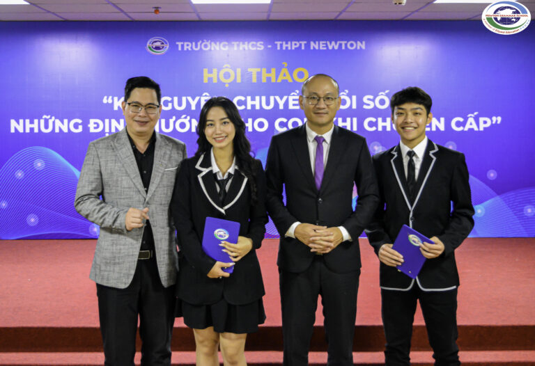 Bốn người đứng trên sân khấu, mỉm cười trước ống kính. Hai người đàn ông mặc vest đứng ở hai bên ngoài, và hai người trẻ mặc áo blazer đen giống nhau và cầm tập tài liệu đứng ở giữa, phía sau họ là phông nền sự kiện màu xanh.